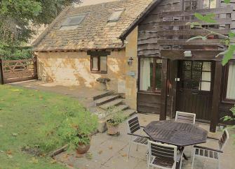 Exterior of a Cotswold honey-stone cottage overlooking a small lawn and patio with a dining table.
