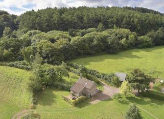 Aerial view of a holiday cottage surrounded by fields beneath a hilltop wood.
