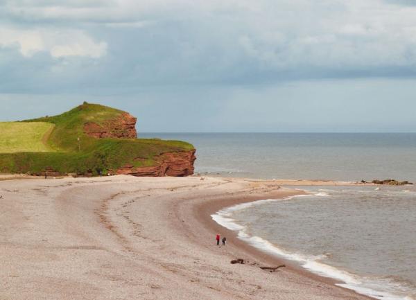 A low sandstone promontory overlooks a curving shingle beach with two walkers walking the wave's edge.