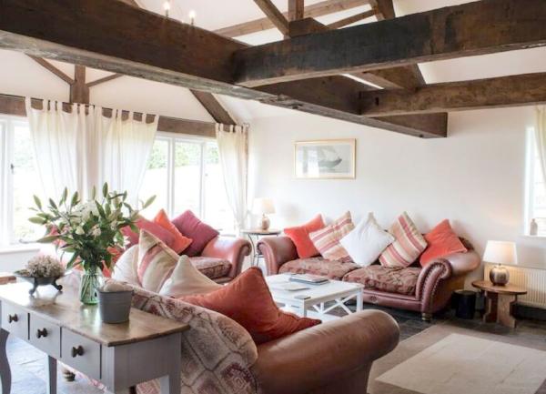A lounge with oak beams and one wall of floor-to-ceiling windows. There are three leather sofas, each with many scatter cushions.