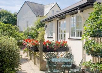 Windows of a little cottage overlook a terrace filled with flowers and shrubs.