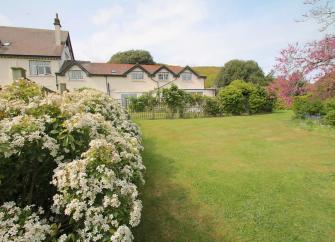 A large Someerset house overlooks a spacious lawn lined by flowering shrubs and trees.