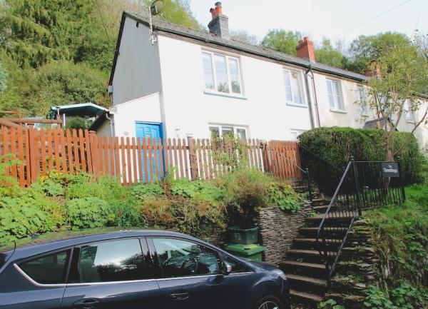A semi-detached countryside cottage with a secure front garden nestles into a tree-covered bank.