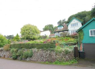 A mature garden slopes up to a large house backed by trees.