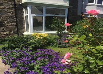 A holiday cottage bay window overlooks a lawn surrounded by flower beds.