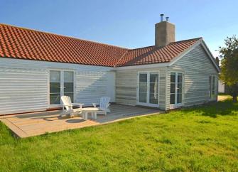 An L-shaped, wood-panelled Essex holiday bungalow with a wooden deck containing a table and sunlounger, all overlooking a large lawn.