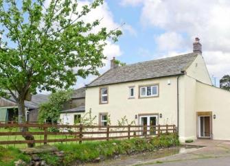 A wood and slate Cotswold holiday cottage overlooks a terrace with a dining table and lawn.