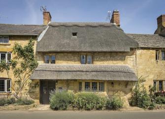 A thatched Cotswold Cottage with a bay window overlooks an empty village street.