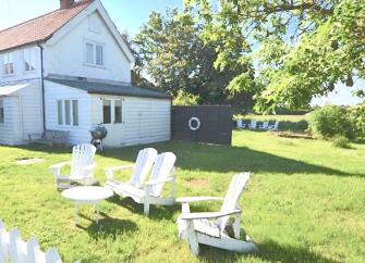A wooden 2-storey Essex holiday cottage overlooks a lawned garden, shaded by tall trees with four sun loungers on the grass.