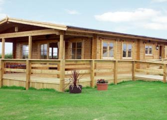 A long eco-friendly log cabin with a covered, wrap-around deck is surrounded by countryside..