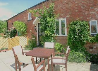 The exterior of a rural holiday cottage has a courtyard garden and a dining table with chairs.
