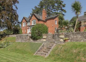 A large brick house backed by tall pine trees overlooks an extensive lawn.