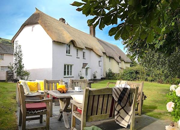 A thatched Winsford holiday cottage with a large lawn and a patio for outdoor dining shaded by trees..