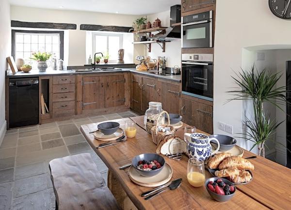 A cottage kitchen with flagstone floors, contemporary appliances integrated within  wooden units and a pine dining table laid for breakfast.