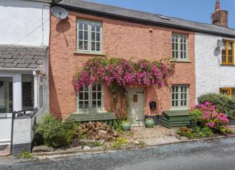 The double-fronted exterior of a rustic, terraced holiday cottage in a Mid Devon village street with a flowery creeper overhanging the front door.