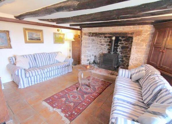 An oak-beamed cottage lounge with a large stone inglenook fireplace containing a wood-burner. Furniture includes two large sofas and a coffee table.