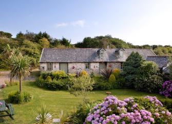 A stone cottage overlooks a lawn bordered by flowering shrubs.