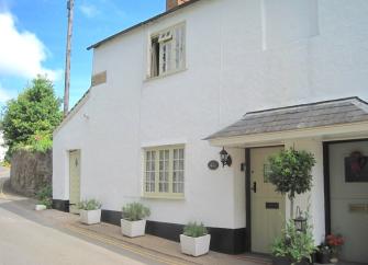 The exterior of a village holiday cottage with a sheltered porch and flower-filled planters on a village street.