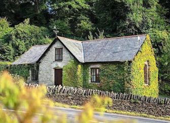 Single-storey, ivy-clad country cottage in front of woodland trees.