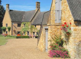 Exterior of a flower-decked Cotswold Manor and adjoining barn facing a shingle drive and lawn.