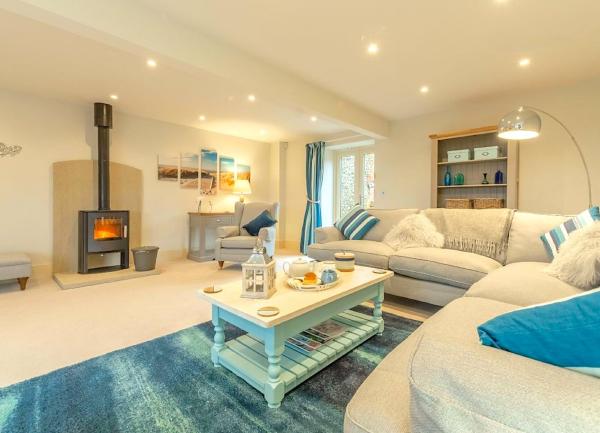 A spacious sitting room with L-shaped sofa, and armchair all warmed by a wood-burner in then fireplace.