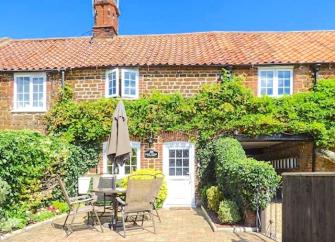 Exterior of a wisteria-clad cottage-front facing a paved front-garden with outdoor dining table, chairs and parasol.