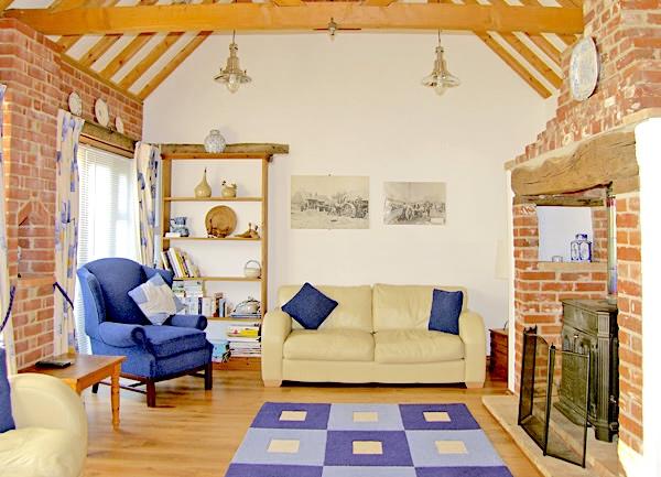 A lounge with pine-beam trusses above a sofa, armchairs and inglenook fireplace with a wood-burner.