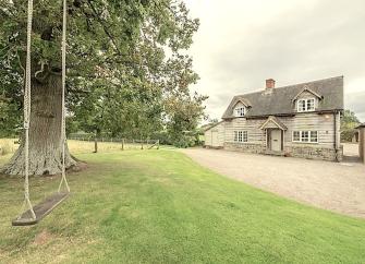 A garden with a garden swing, trees and a wide drive are overlooked by a weather-boarded 2-storey Herefordshire cottage.