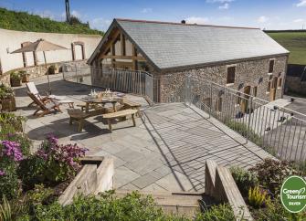 Larger raised and paved terrace with picnic tables and railings overlooks a stone barn conversion and fields.
