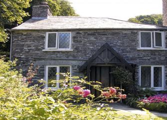 A double-fronted stone cottage overlooks a sunny lawn edged with flowers.