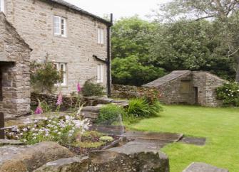 A stone cottage with a large porch overlooks a walled lawn and flower beds.
