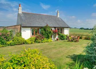 A holiday bungalow with a flowerbeds and a lawn surrounded by open fields.