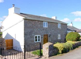 A double-fronted stone cottage with a hedged-in lawn to the front and a gated driveway for cars.