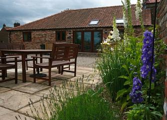 Single-storey barn conversion overlooks a spacious courtyard with dining furniture bordered by flower beds.