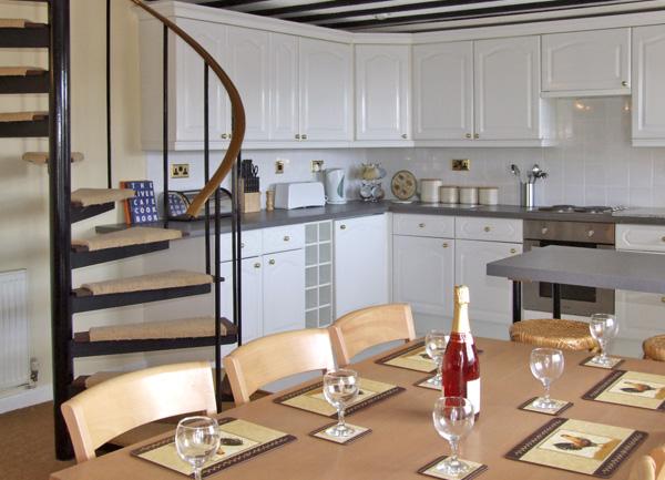Behind a wooden dining table laid for tea is a kitchen with fitted units, oven and sink.
