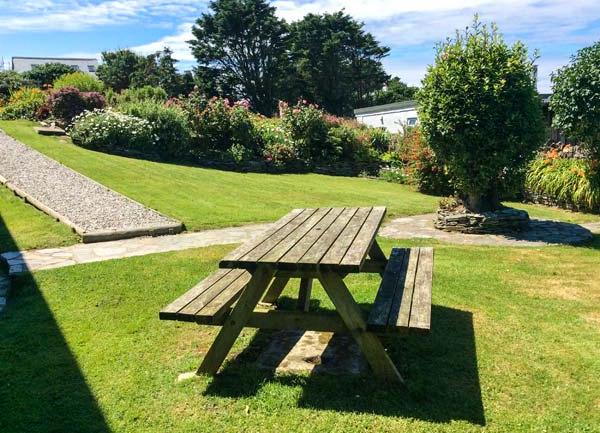 A large lawn with a picnic bench is bordered by trees and shrubs with a sea view beyond.