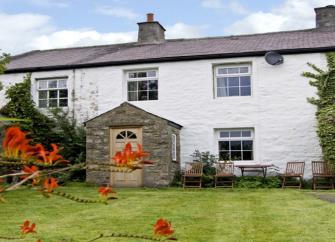 A whitewashed cottage with a stone-built porch overlooks a lawn lined by mature shrubs and flowers.