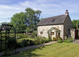 Peak District stone barn conversion with a wisteria-clad pergola over the front door is surrounded by large lawns either side of a garden path.