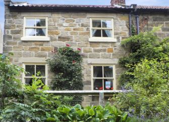 A semi-dtached, stone-buit North Yorkshire holiday cottage sits behind afront garden with shrubs and roses.