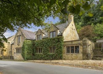 A 2-storey, Cotswold honeystone cottage backed by tall trees.