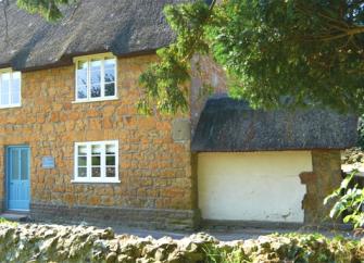 A honey-stone, thatched country cottage with a walled front garden.