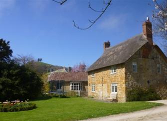 Exterior of a stone-built thatched village cottage overlooking a large lawn with a flowerbed.