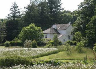A country cottage on the edge of a wood surrounded by well-kept lawns and flower beds.