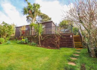 A single-storey timber lodge sits above a raised deck above a large, shrub-lined lawn