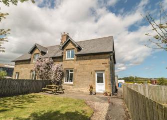 A semi-detached stone farm cottage with a lawn and picnic table.