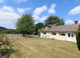 A single-storey Dorset holiday cottage overlooks a large lawn surrounded by a wooden fence, trees and rose bushes.