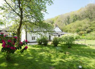 An old mill house beneth a wooded hill surrounded by gardens.
