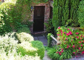 A stout wooden door in an ivy-clad stone-built porch surrounded by shrubs and flowers.