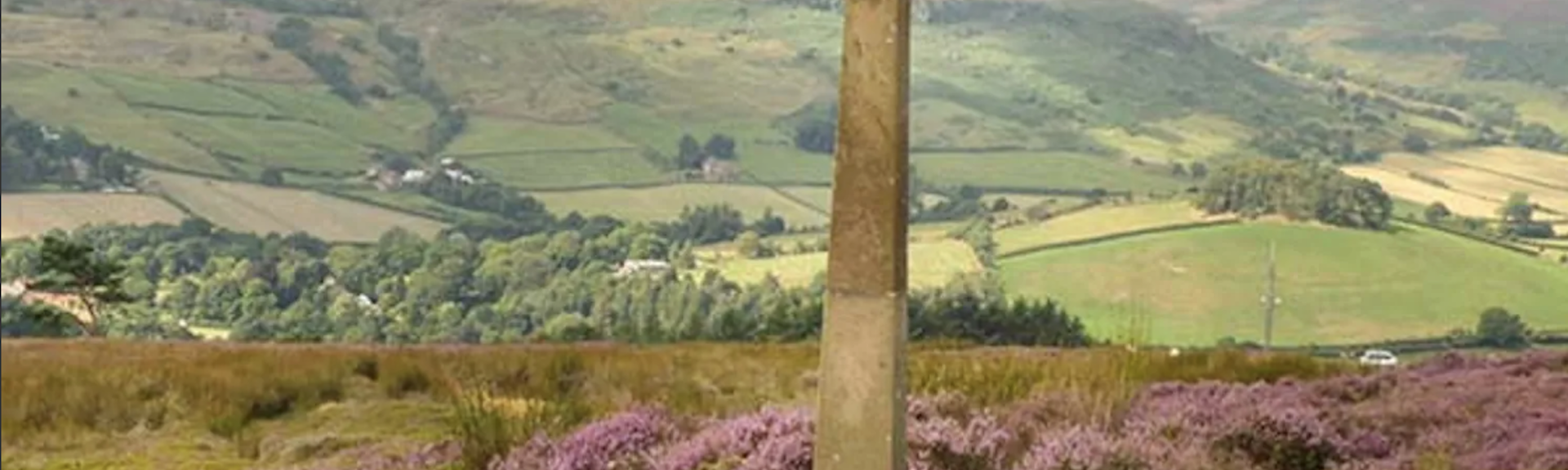 a roadside celtic cross on th purple heather covered moorland with far reaching views across a wide valley t