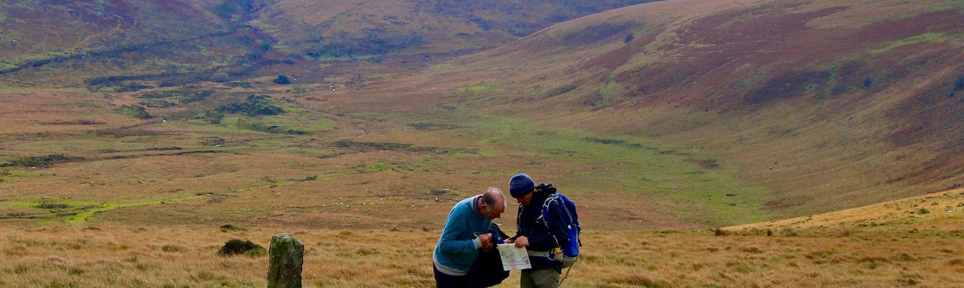 Two Dartmoor walkers study maps next to a standing stone in an open expanse of of moorland with a large hilly, moorland Tor in the background